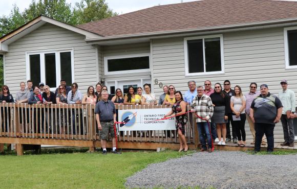 OFNTSC staff and visitors stand on the deck in front of the new office in Atikameksheng Anishnawbek. Board member Paul Schisler and Executive Director Melanie Debassige hold a red ribbon and scissors ready to cut the ribbon signifying the official opening.