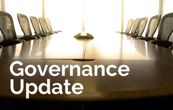 A dark brown wood boardroom table with black chairs and the words "Governance Update" superimposed in the bottom left corner