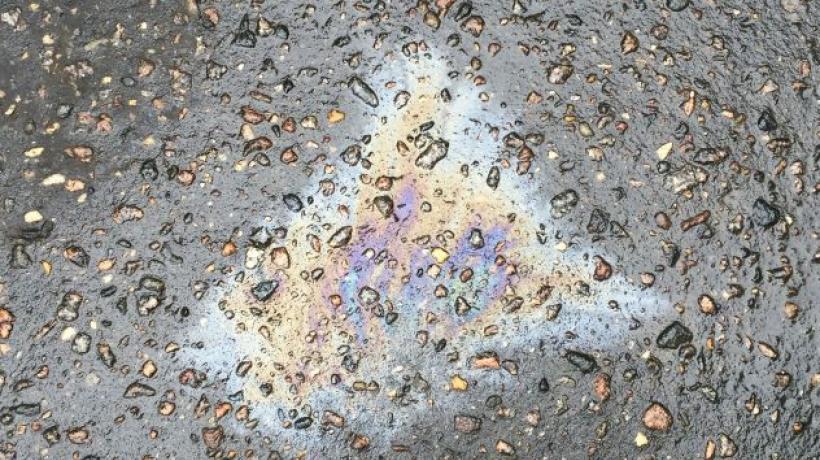 An image of an oily fuel spill on pavement, which creates a rainbow like-hue on the black stoney ground. 